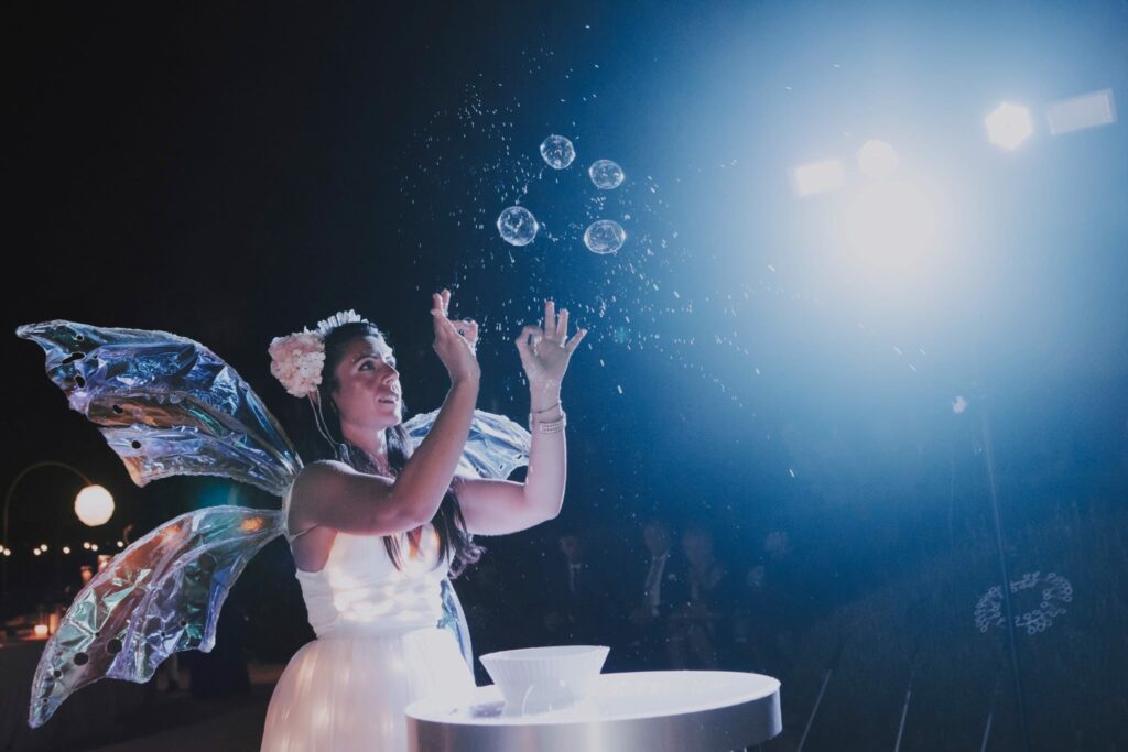 Uno spettacolo di bolle di sapone con abiti luminosi può essere un’esperienza visivamente affascinante e divertente. Questo tipo di spettacolo combina la magia delle bolle di sapone con l’aggiunta di abiti o costumi che emettono luce o che sono decorati con luci luminose. Immagina un artista delle bolle di sapone che indossa un abito che si illumina al buio o che ha luci integrate al suo interno. Mentre soffia bolle di sapone, queste potrebbero riflettersi e trasmettere la luce emessa dall’abito, creando un effetto straordinario. Le bolle potrebbero sembrare sospese nel vuoto, come sfere luminose che danzano nell’aria.